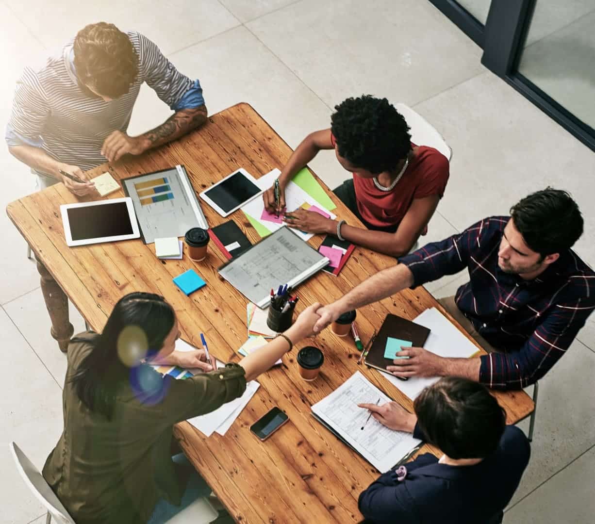 Five people, including a digital consultant, sit around a wooden table with laptops, tablets, papers, and coffee cups, collaborating and shaking hands in a modern office setting.