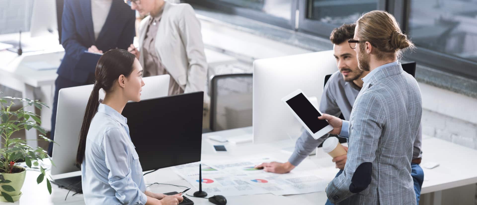 Three people are having a discussion in a modern office; a digital consultant holds a tablet, while another sits at a desk with paperwork and computer monitors. Two more people talk in the background.