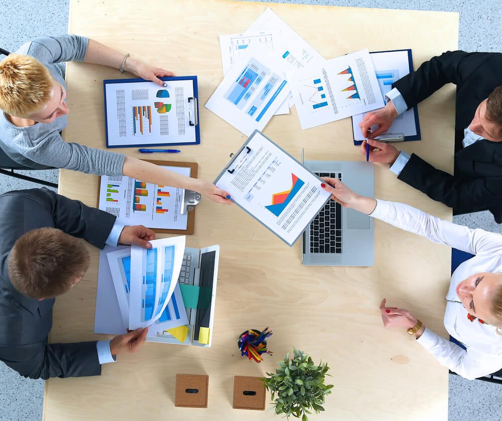 Four people sit around a table reviewing documents with charts and graphs, laptops and papers spread out for a business meeting on White Label SEO Services for Agencies.