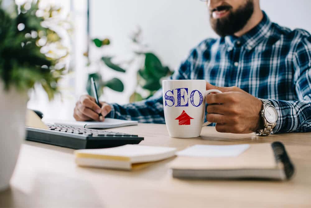Man in a plaid shirt holding a mug labeled "SEO" while writing in a notebook at a desk, planning private label SEO services with a calculator, notepads, and a plant in the background.
